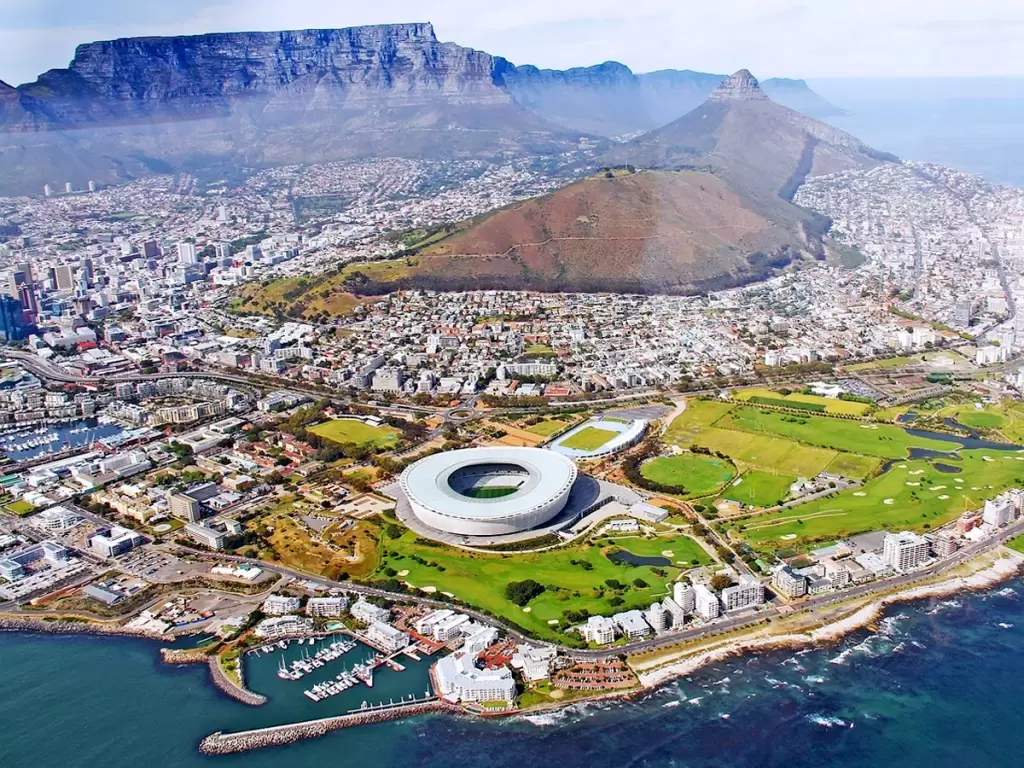 Aerial view of Cape Town, showcasing the iconic Table Mountain, Cape Town Stadium, and the vibrant waterfront, surrounded by the vast Atlantic Ocean and stunning cityscape.