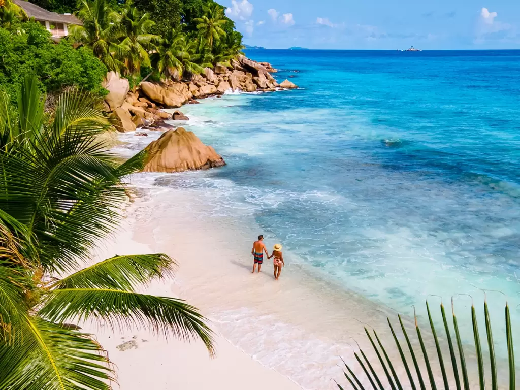 A couple walking along the white sand beach of Seychelles' Anse Source D'Argent, with crystal-clear turquoise waters and lush palm trees.