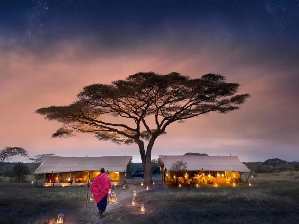Luxury African safari tents lit, with a Maasai guide in a remote Serengeti camp.
