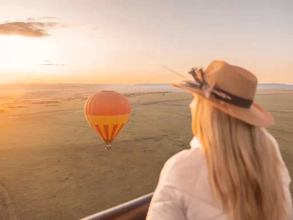 A woman viewing a hot air balloon while drifting over golden plains at sunrise during a luxury Serengeti safari in Africa.
